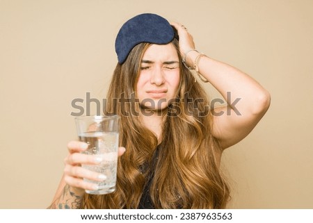 Ill young woman with a sleep mask drinking water with medicine after waking up in the morning feeling sick with a headache and hangover