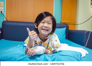 Illness Asian Child Smiling Happily And Showing Thumb Up Hand Sign. Girl Admitted In Hospital While Saline Intravenous (IV) On Hand. Health Care Stories. 