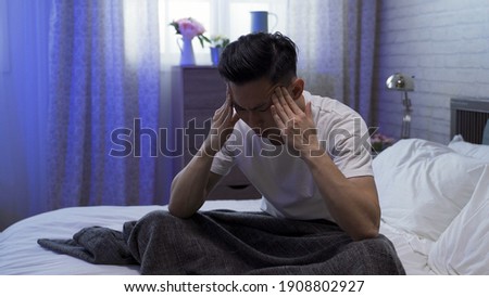 illness asian guy sitting on bed is having bad headache, feeling stressful and rubbing his temples with both hands.