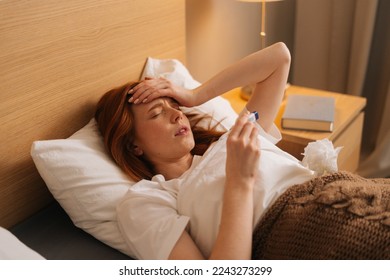 Illness tired woman touching forehead after using thermometer to checking body temperature and take medicine lying under blanket on bed. Sick female with cold lying in bed with high fever in bedroom.