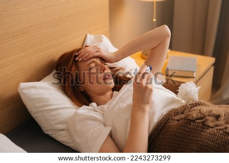 Illness tired woman touching forehead after using thermometer to checking body temperature and take medicine lying under blanket on bed. Sick female with cold lying in bed with high fever in bedroom.