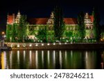 Illuminated, brick, historic building of National Museum in Wroclaw overgrown with ivy at night with reflection in the Odra river.