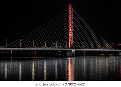 Illuminated cable-stayed bridge spanning across dark water at night in Hangzhou, China, featuring a striking red central tower and symmetrical cable design with reflections.