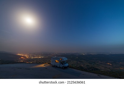 Illuminated Camper Van On Road Side In Beautiful Night Landscape. Moon In The Sky Over Valley And Villages, Unique Highlands And Hill Range In Italy, Alternative Vanlife Vacation Concept.