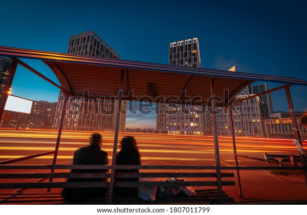 An illuminated city street at the\
night. The bus stop with silhouettes of a couple, sitting on the\
bench, in front of bright light trails, from moving\
cars.