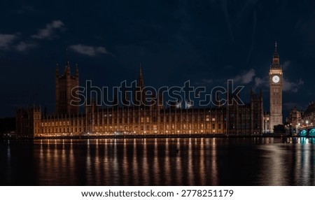Illuminated clock tower and long gothic building stand beside river under dark sky. Warm lights reflect on water forming clear architectural night scene.