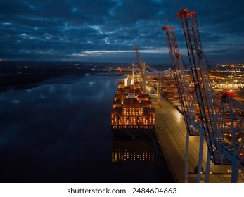 Illuminated container ship and Southampton Docks at dusk. High latitude aerial view of the port cranes and stern of the container ship with lights reflection on the sea. Calm cloudy night. - Powered by Shutterstock