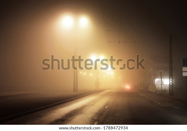 Illuminated empty asphalt road in a fog at\
night. Dark urban scene, cityscape. Industrial zone in Riga,\
Latvia. Dangerous driving, speed, freedom, concept\
image