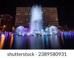 An illuminated fountain with vibrant colors situated in front of a grand hotel overlooking a lake, illuminated in the night