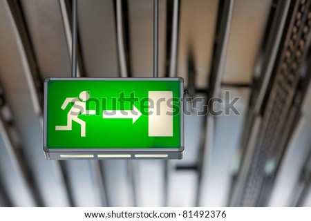Illuminated green exit sign suspended from the ceiling in a public transportation facility. Signage consists of a human figure running and an arrow pointing at a door.