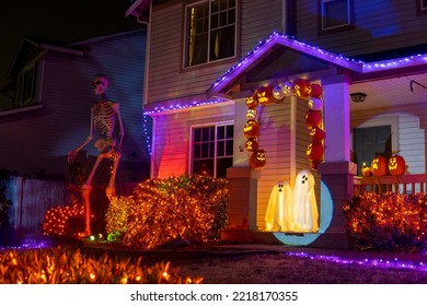 Illuminated Halloween house outdoor decorations with orange and purple garlands, skeleton, pumpkins and ghosts near the house porch