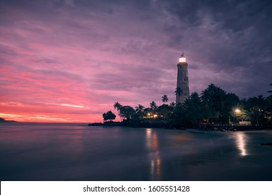 Illuminated lighthouse in the middle of palm trees against dramatic sky. South coast of Sri Lanka at colorful sunrise. 
