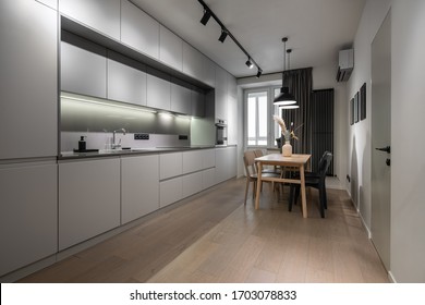 Illuminated modern kitchen with light walls and a parquet. There are gray lockers, sink with faucet, stove, oven, wooden table with vase and chairs, window with curtain, different lamps, door. - Shutterstock ID 1703078833