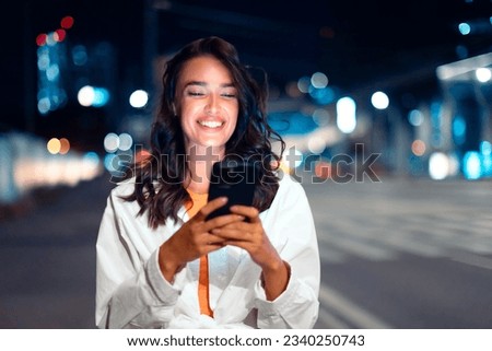 Illuminated promenade. Happy caucasian woman texting via smartphone, walking in city with night illumination on background. Cheerful lady having promenade in megalopolis