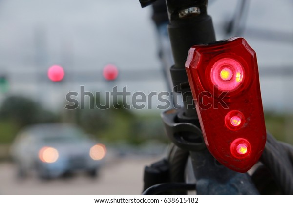 Illuminated Red Rear Bicycle Light Close Up Frame\
Right with Red Traffic Lights and Cars with Headlights Moving\
Making a Right Turn in the Background against Gray Cloudy Overcast\
Sky at Dusk