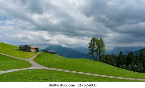 illuminated wooden alpine farmhouse with view to the steep mountains from Bregenz forest, under dark clouds from thunderstorm. by the road through green meadows with trees