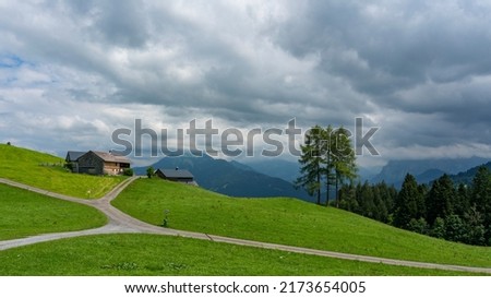 illuminated wooden alpine farmhouse with view to the steep mountains from Bregenz forest, under dark clouds from thunderstorm. by the road through green meadows with trees