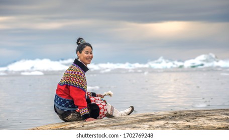 Ilulissat, Greenland - July 11, 2018: Young Inuit Woman In Traditional Clothing Posing For Photos On The Shore Of The Ocean, Nearby By Small Greenlandish Village.