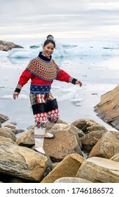 Ilulissat, Greenland - July 11, 2018: Young Inuit Woman In Traditional Clothing Posing For Photos On The Shore Of The Ocean, Nearby By Small Greenlandish Village.