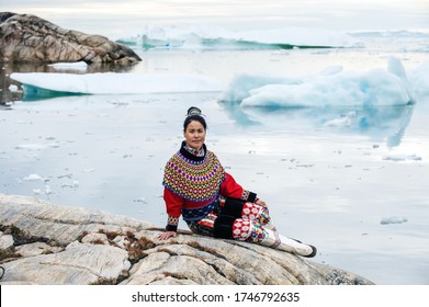 Ilulissat, Greenland - July 11, 2018: Young Inuit Woman In Traditional Clothing Posing For Photos On The Shore Of The Ocean, Nearby By Small Greenlandish Village.