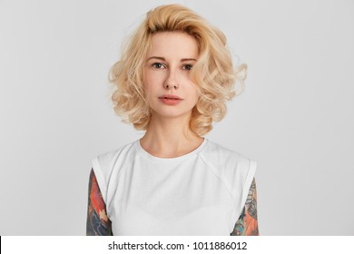 Horizontales Porträt eines jungen blonden Mädchens, das ein weißes lässiges T-Shirt trägt, mit gepiercter Nase, aufrecht steht und nachdenkt. Ernst. Isoliert auf weißer Wand