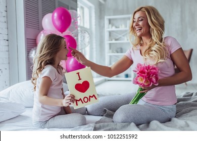 I love you, mom! Attractive young woman with little cute girl are sitting on bed and spending time together at home. Mom is receiving presents from daughter on Mother's Day. Happy family concept.