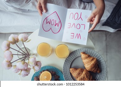 Wir lieben dich, Mama! Zugeschnittenes Bild einer attraktiven jungen Frau sitzt zu Hause auf dem Bett. Blick von oben auf das Frühstück im Bett mit einer Kinderpostkarte am Muttertag.
