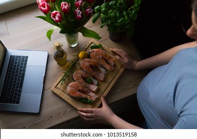 La niña embarazada está viendo el curso en línea y preparando salmón con romero y limón para el almuerzo. En la mesa hay flores de tulipán frescas. Vista superior