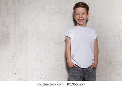 Cute little boy in white T-shirt posing in front of grunge concrete wall. Portrait of fashionable male child. Smiling boy posing, gray wall on background. Concept of children style and fashion.