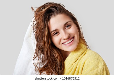 濡れた髪の素敵な女性、シャワーを浴び、タオルで頭を乾かし、入浴後に喜んで、黄色いバスローブを着て、白い背景にポーズをとり、明るい表情をしています