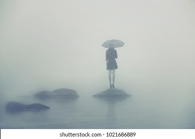 Girl with umbrella standing on a rock in the middle of the water. Alone woman in the fog