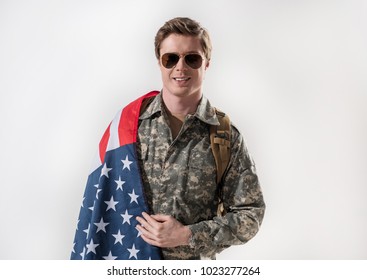 Waist up portrait of confident military young man looking at camera and smiling. American Flag is lying on his shoulder and he is having rucksack behind his back. Isolated on background