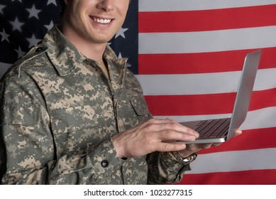 Joyful young military man smiling and having device in his hands while typing. Flag on background