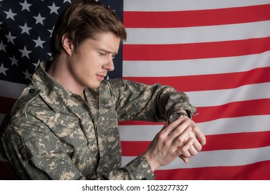 Thoughtful male soldier standing on background of the flag. He is looking at his watch and frowning