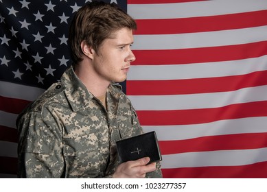 Tranquil young military man standing and holding the Bible in his hand with serene look. USA Flag on background. Copy space in right side