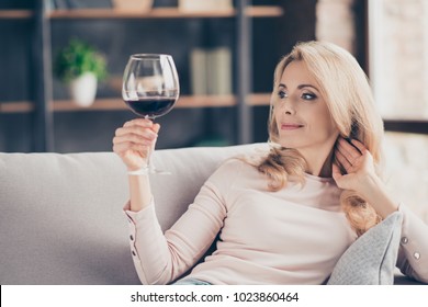 Portrait of pretty, charming, attractive, stylish connoisseur woman sitting on couch having raised glass with red wine in hand, examine, taste beverage