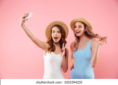 Two brunette and blonde women in one-piece swimsuits and straw hats smiling and taking selfie on smartphone isolated over pink background