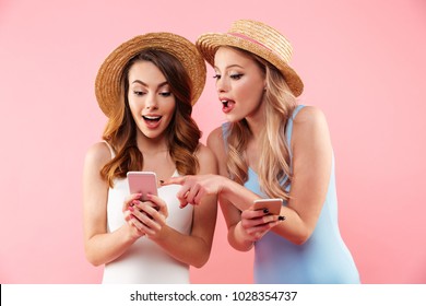 Bild von zwei prächtigen Frauen, die einteilige Badeanzüge und Strohhüte tragen, die im Internet surfen oder Mobiltelefone verwenden, isoliert auf rosa Hintergrund
