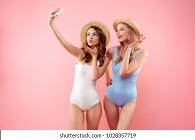 Two brunette and blonde women in one-piece colorful swimsuits and straw hats smiling and taking selfie on cell phone isolated over pink background