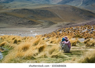 若い女性旅行者が湖岸に座って、ボリビアのアルティプラノで双眼鏡を使ってフラミンゴを見ています。未舗装の道路のある火山の風景が背景にある