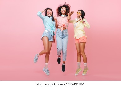 Retrato completo de una entusiasta chica africana con zapatos negros posando entre amigos internacionales en un estudio rosa. Mujer asiática deportiva con camisa azul saltando cerca de damas elegantes.