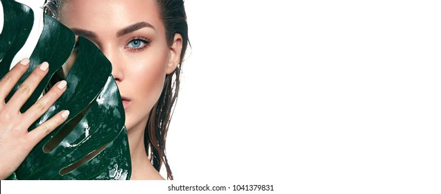 A beautiful young girl in the studio on a white background with wet skin and wet hair holds a large green tropical leaf in hands and covers a part of her face.Fashion, beauty, make-up, cosmetics.