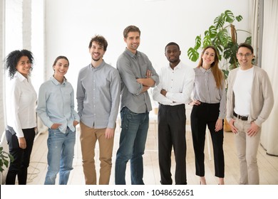 カメラの笑顔を見ている若い幸せな多民族の専門家または会社のスタッフ、一緒に立っている多様なビジネスマンの多民族グループ、オフィスでポーズをとる従業員、成功したチームのポートレート