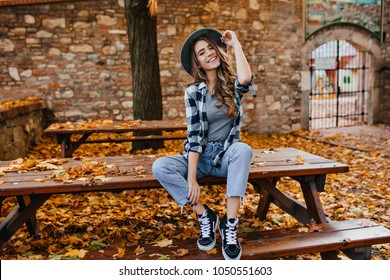 Mujer joven despreocupada en pantalones de moda vintage sentado en la mesa en el parque y riendo. Linda chica rizada de buen humor posando en día de otoño, disfrutando del buen tiempo.