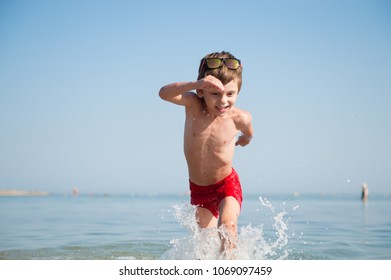 Anak kecil yang sehat, lucu, dan bahagia mengenakan kacamata hitam dan celana pendek merah berlari di air laut dengan cipratan air di liburan musim panas