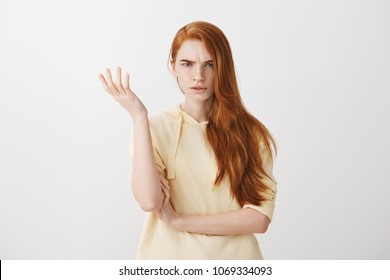De qué diablos estás hablando, tonterías. Foto de estudio de mujer frustrada con cabello rojo gesticulando con la palma levantada, frunciendo el ceño, disgustada y confundida con una pregunta tonta sobre la pared gris