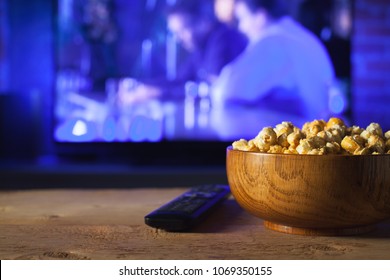A wooden bowl of popcorn and remote control in the background the TV works. Evening cozy watching a movie or TV series at home
