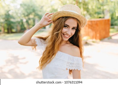 Nahaufnahmeporträt der inspirierten jungen Dame mit den schönen Augen, die Strohhut halten. Romantisches blondes Mädchen mit blasser Haut, die auf unscharfem Hintergrund aufwirft und lächelt.
