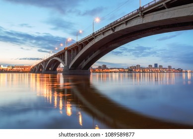 Die Brücke über den Jenissei leuchtet mit Abendlichtern und spiegelt sich in der Wasseroberfläche während des Sonnenuntergangs in Krasnojarsk, Russland. Ein Modell des sowjetischen Brückenbaus in den 1950er und 1960er Jahren.