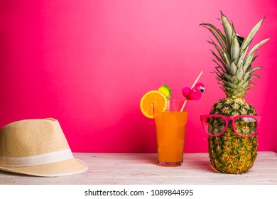 Stijlvolle ananas met zonnebril, strohoed en limonade met sinaasappel en tubulle met flamingo aan de witte tafel over roze achtergrond. Zomer compositie